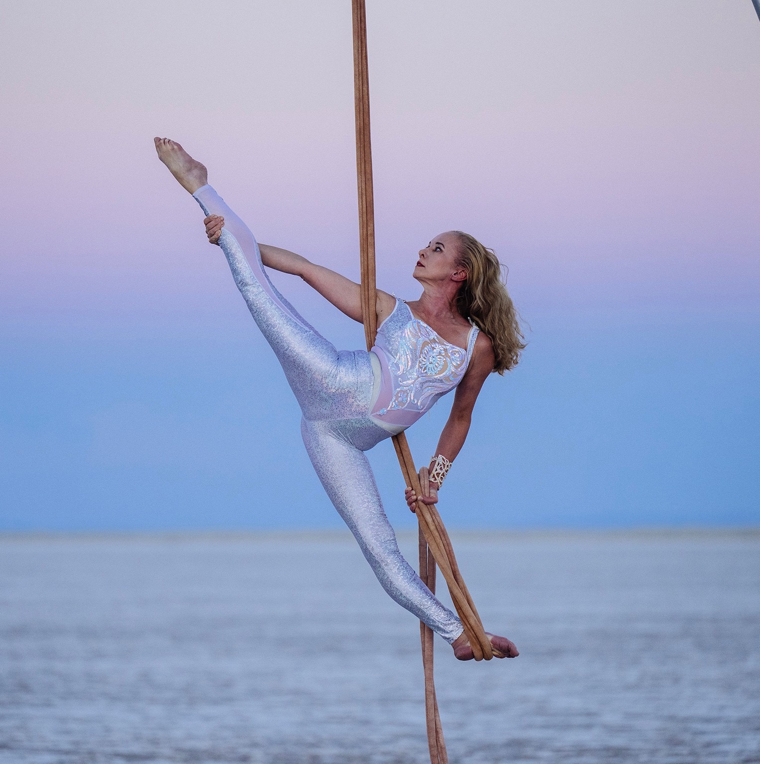 Location shot. A white woman with long blonde hair wearing a white spandex unitard. She's on a vertical rope holding a split-legged pose from an aerial act, behind her is the ocean and a dusky sky.