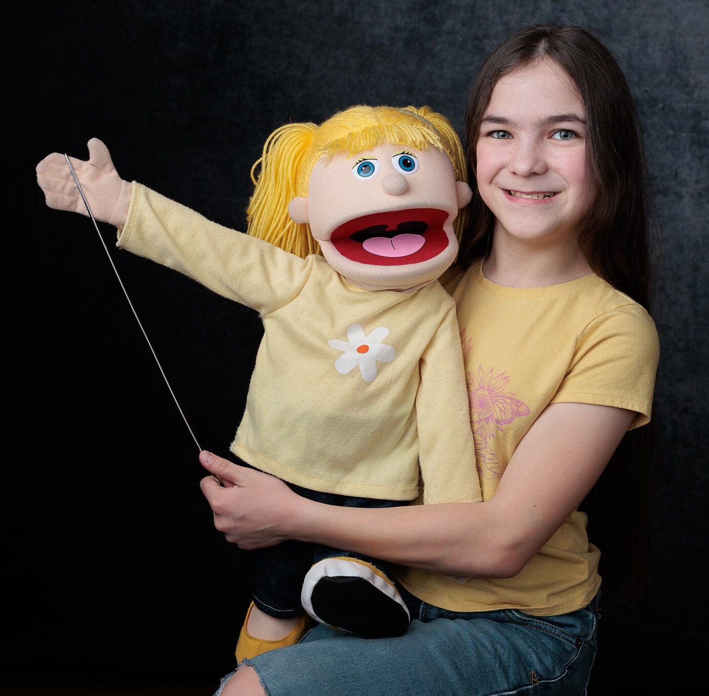A girl in a yellow t-shirt, smiling, holds a blonde-haired, blue-eyed puppet girl on her lap. They are both looking at the camera and smiling.