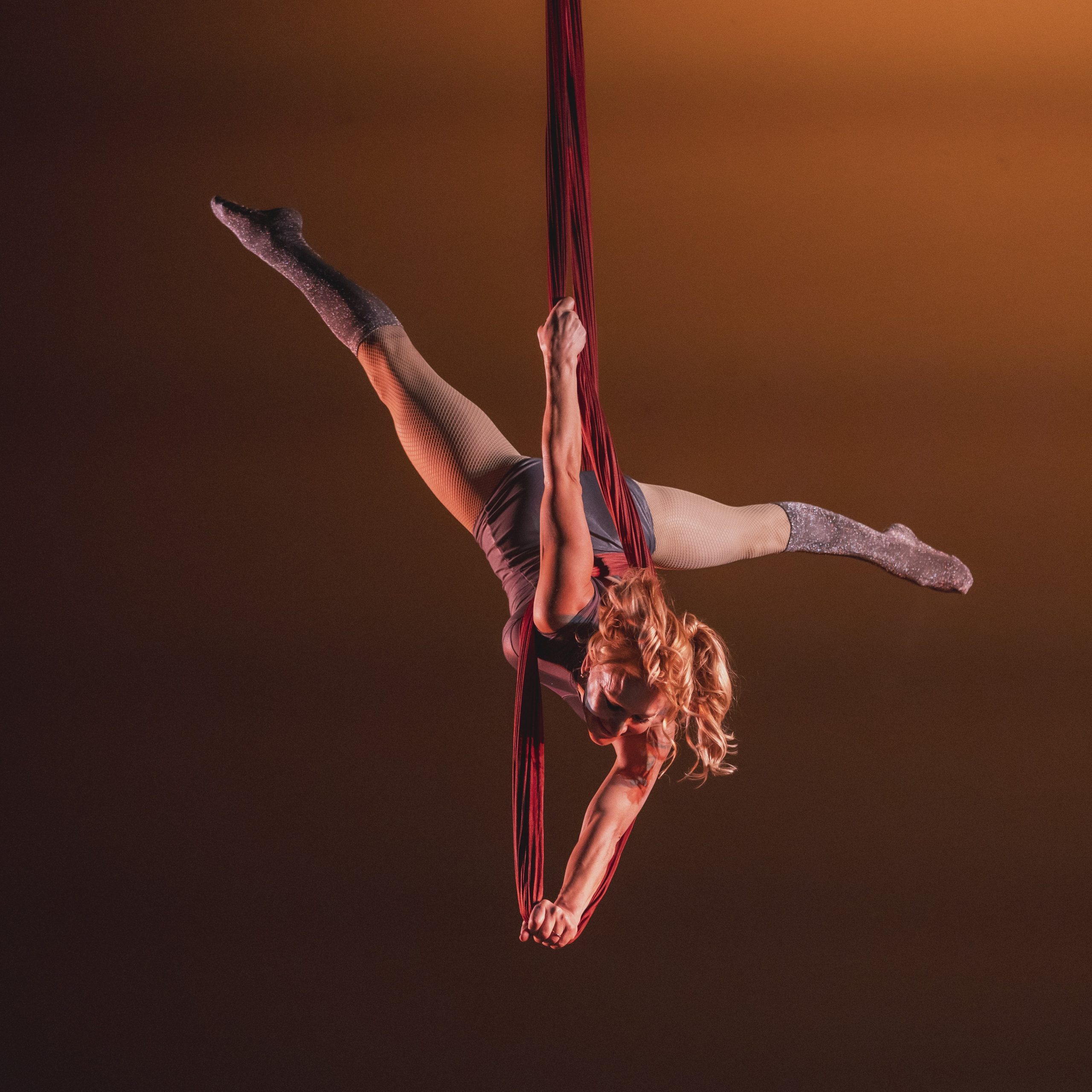A white woman with blond hair performing an aerial silks routine against a tawny brown background.