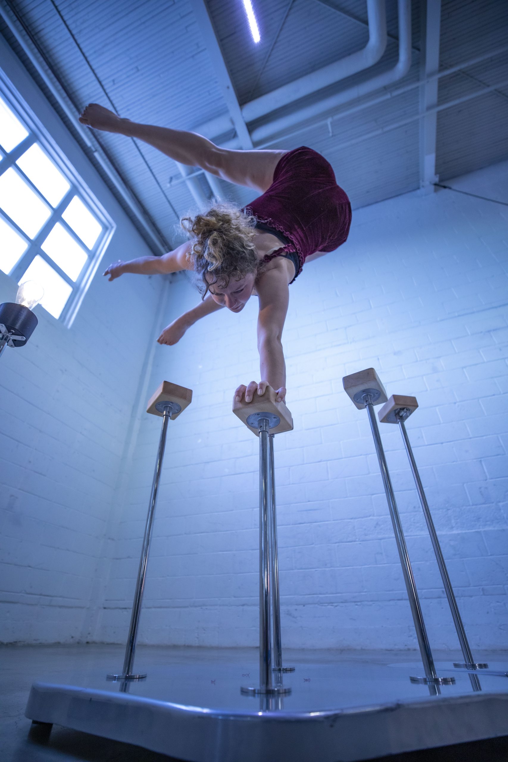Photographed from the floor, the camera looks up underneath a blonde, white woman balancing on one hand on a handstand cane. She's in a coolly-lit, white painted wall warehouse space.